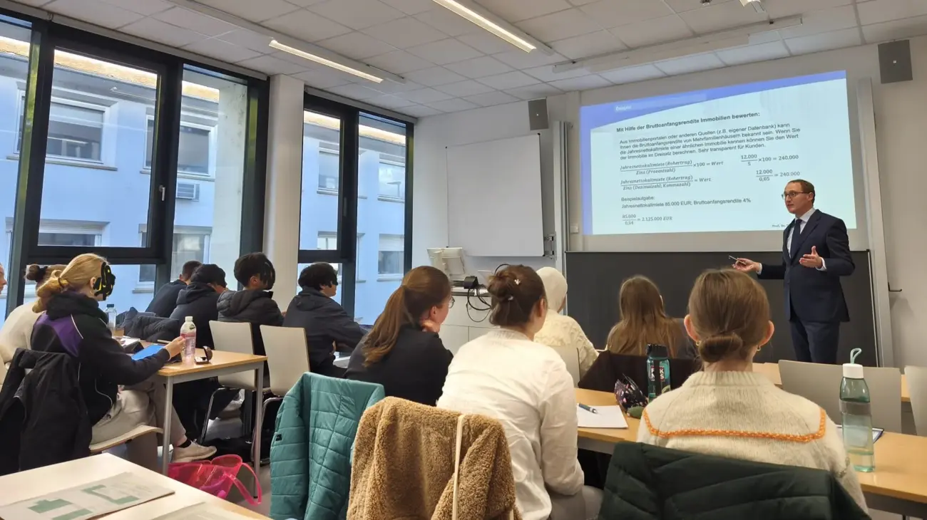 Blick von hinten in einen Hörsaal mit Schüler:innen, die dem Vortrag eines Professors folgen.