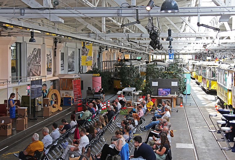 weiter Blick in die Halle des Museums mit historischen Straßenbahnen und Publikum und Rednerin
