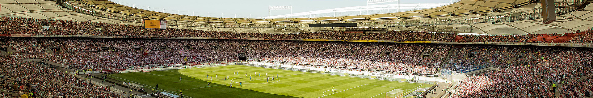 Das Bild zeigt ein großes, modernes Fußballstadion, das mit Zuschauern gefüllt ist. Die Architektur umfasst ein teilweise überdachtes Dach, und das Spielfeld ist grün und gepflegt. Auf dem Feld sind Spieler zu sehen, die gerade ein Spiel austragen. Weitere Details: Die Tribünen sind voll besetzt und sorgen für eine lebendige Atmosphäre. Entlang des Spielfeldrands befinden sich Werbebanner. Es sind verschiedene Sitzbereiche erkennbar, darunter auch Abschnitte für unterschiedliche Fangruppen.