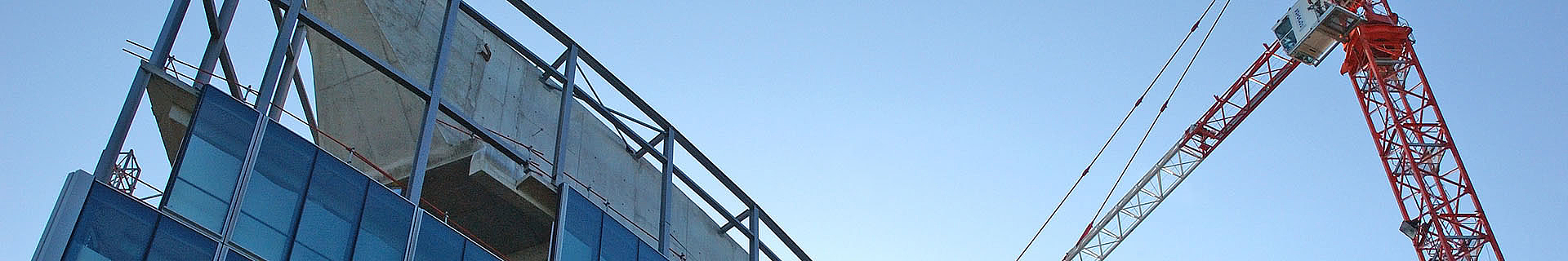 Das Bild zeigt eine Baustelle mit einem teilweise errichteten Gebäude und einem großen rot-weißen Baukran. Die unteren Stockwerke des Gebäudes sind bereits mit Glasfenstern versehen, die oberen Stockwerke sind noch im Rohbau mit sichtbarem Beton und Stahlgerüst. Der Kran ragt hoch über das Gebäude und deutet auf aktive Bauarbeiten hin. 
