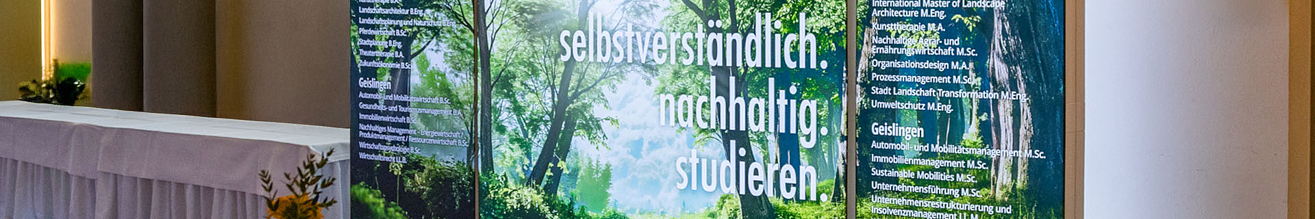 Sliderbild Messe. Das Bild zeigt einen Auschnitt des Messestandes der HfWU. Darauf sind die Worte "selbstverständlich. nachhaltig. studieren" auf einem Hintergrund mit Wald zu sehen.