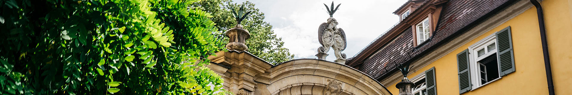 Das Bild zeigt den oberen Teil eines historischen Gebäudes mit einem kunstvoll gestalteten Steinbogen. Der Bogen ist mit dekorativen Elementen verziert, darunter zwei Skulpturen mit vogelähnlichen Figuren auf der Oberseite. Links vom Bogen ist üppiges grünes Laub von Bäumen oder Pflanzen zu sehen. Rechts erkennt man einen Teil eines gelben Gebäudes mit grünen Fensterläden und einem geöffneten Fenster. Der Himmel im Hintergrund ist teilweise bewölkt.