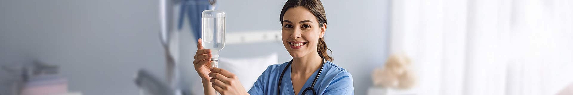 Das Bild zeigt eine Person in medizinischer Kleidung (Scrubs), vermutlich eine medizinische Fachkraft, die einen Infusionsbeutel hält. Das Gesicht der Person ist unscharf dargestellt. Im Hintergrund erkennt man eine klinische Umgebung mit medizinischen Geräten und Materialien – typisch für ein Krankenhaus oder eine Praxis.