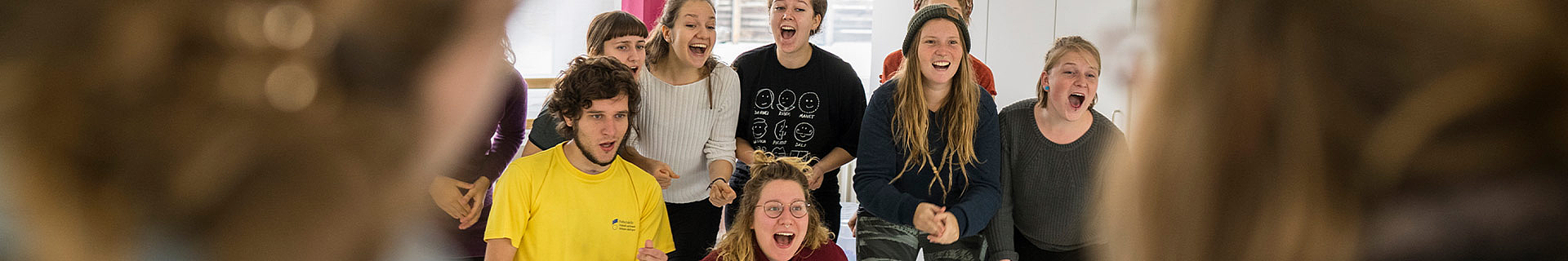 Das Bild zeigt eine Gruppe von Menschen in einem Seminarraum. Es sind mindestens acht Personen zu sehen – einige stehen, andere sitzen, und manche sind teilweise von anderen verdeckt. Die Gesichter sind aus Datenschutzgründen unscharf dargestellt. Im Hintergrund erkennt man Fenster und weiße Wände.