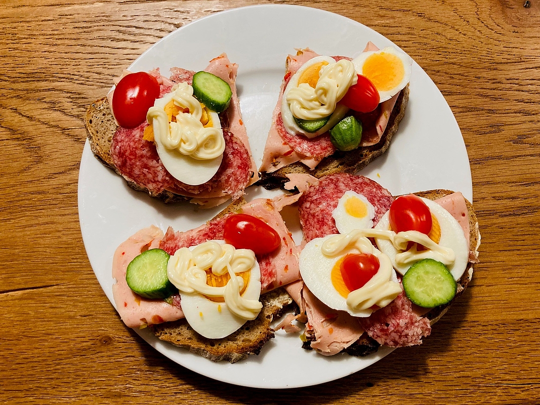 Teller auf einem Holztisch mit vier "Schnittchen", also belegten Broten mit Wurst, Salami, Ei, Gemüsedekoration und Mayonnaise