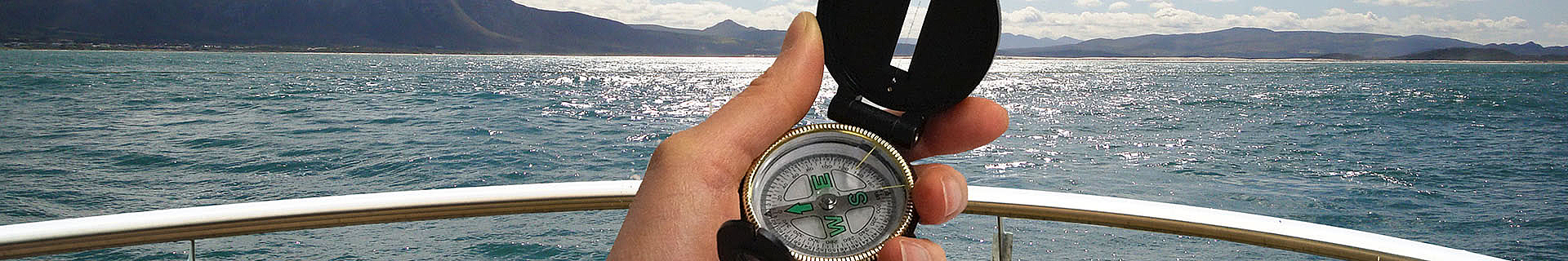 Das Bild zeigt eine Hand, die einen Kompass auf einem Boot hält. Im Hintergrund erstreckt sich das offene Meer, mit Bergen in der Ferne und einem teils bewölkten Himmel darüber. Der Kompass zeigt nach Norden, und im Vordergrund ist das Geländer des Boots zu sehen. Das Bild vermittelt ein Gefühl von Orientierung, Navigation und Abenteuer. 