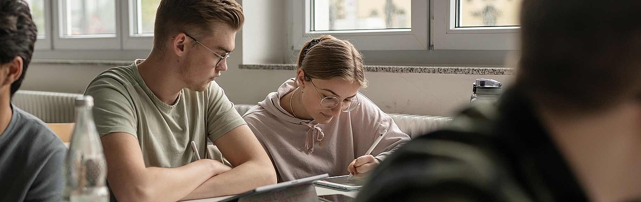 Das Bild zeigt zwei sitzende Studierende in einem Vorlesungsraum, die an ihren Tablets arbeiten. 