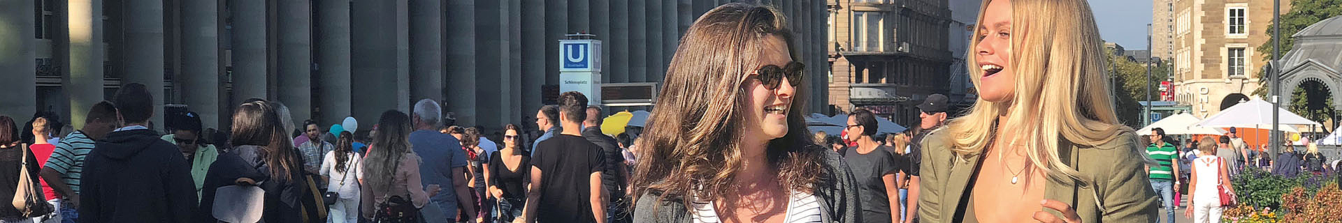 Das Bild zeigt eine belebte Szene im Freien mit vielen Menschen, die herumlaufen oder stehen. Im Hintergrund sind große Säulen zu sehen, vermutlich Teil eines bedeutenden Gebäudes oder einer öffentlichen Struktur. Ein Schild mit dem Buchstaben „U“ weist auf einen U-Bahn-Eingang hin. 