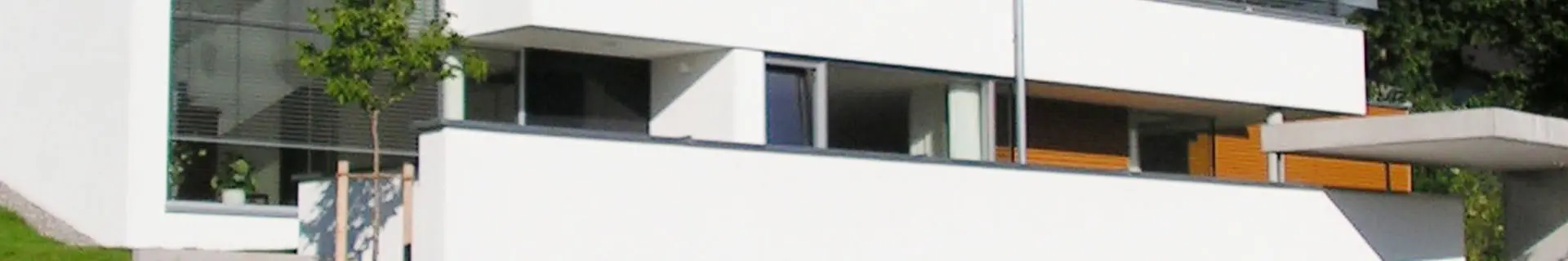 Das Bild zeigt ein modernes, minimalistisches weißes Gebäude mit großen Fenstern und einem Flachdach. Vor dem Gebäude steht ein kleiner Baum, und ein Teil der Architektur umfasst ein Vordach oder eine Überdachung. Das Design zeichnet sich durch klare Linien und eine Kombination aus Glasflächen und festen Wandstrukturen aus.