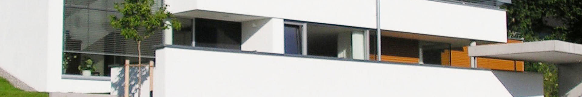 Das Bild zeigt ein modernes, minimalistisches weißes Gebäude mit großen Fenstern und einem Flachdach. Vor dem Gebäude steht ein kleiner Baum, und ein Teil der Architektur umfasst ein Vordach oder eine Überdachung. Das Design zeichnet sich durch klare Linien und eine Kombination aus Glasflächen und festen Wandstrukturen aus.