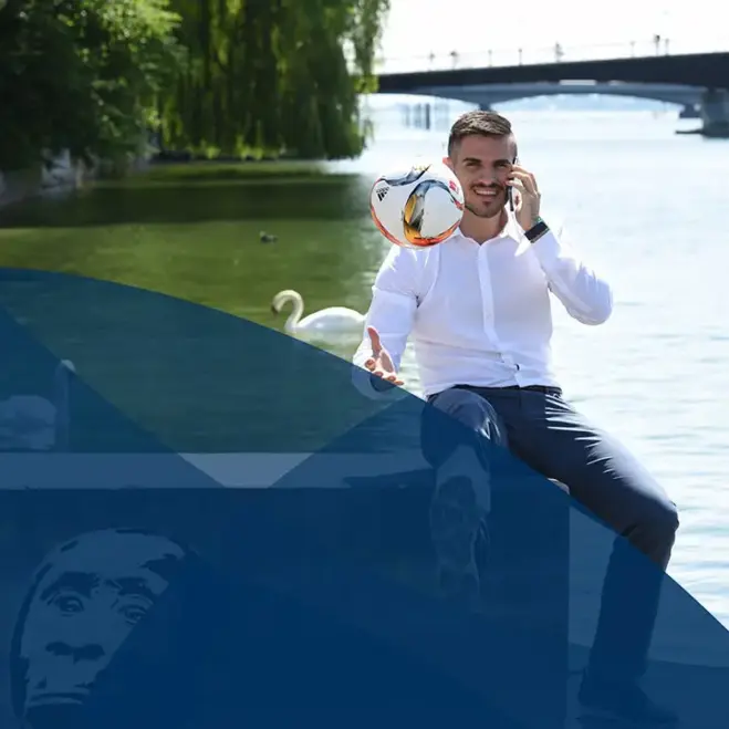 Studierender mit einem weißen Hemd sitzt auf einer Mauer vor einem See und spielt einen Fußball in die Luft