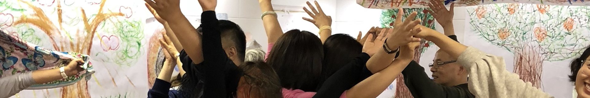 Das Foto zeigt eine Gruppe von Menschen, die vor einer Wand stehen, die mit bunten Zeichnungen von Bäumen und Blumen verziert ist. Sie alle haben ihre Hände erhoben und scheinen an einer gemeinsamen Aktivität beteiligt zu sein, möglicherweise halten sie ein großes Stück Stoff oder ein Kunstwerk hoch. Auf dem Bild sind keine Texte oder Schriftzeichen zu sehen.