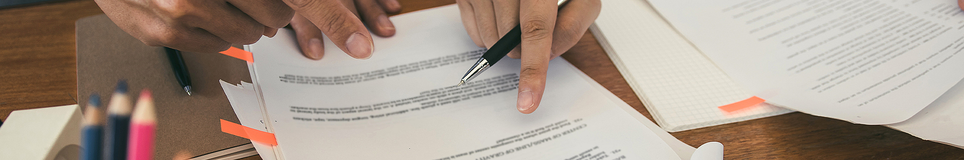 Das Bild zeigt eine Nahaufnahme von Händen, die mit Dokumenten auf einem Holztisch arbeiten. Eine Hand hält einen Stift und zeigt auf einen Text auf einem Blatt Papier, während die andere Hand ein weiteres Dokument hält oder anpasst. Auf dem Tisch liegen mehrere Blätter mit gedrucktem Text, einige davon mit orangefarbenen Markierungen. Im Hintergrund sind auch Buntstifte zu sehen. Die Szene wirkt wie eine Arbeitssituation oder ein Lernumfeld, jedoch sind keine spezifischen Aufgaben oder mathematischen Inhalte erkennbar.