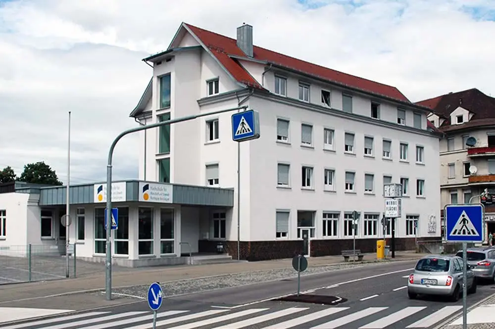 Das Bild zeigt das Campusgebäude Ba37 der HfWU. Es liegt an der Bahnhofstrasse 37 in 73321 Geislingen an der Steige. Es ist ein mehrstöckiges Gebäude mit einem roten Dach und weißen Außenwänden. Das Gebäude hat viele Fenster pro Etage und große Glasfenster im Erdgeschoss. Es befindet sich an einer Straßenkreuzung mit Zebrastreifen und Verkehrsschildern, die auf den Fußgängerüberweg hinweisen. Hier befindet sich auch der Eingang zum Gebäude.