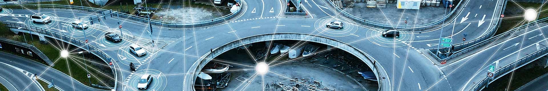 Das Bild zeigt eine Luftaufnahme eines komplexen Straßenkreuzes mit mehreren Ebenen und Fahrspuren. Auf den Straßen sind mehrere Autos und ein Motorrad unterwegs. Über das Bild sind digitale Grafiken gelegt, die Kommunikationssignale oder Vernetzung zwischen den Fahrzeugen darstellen – ein Hinweis auf ein intelligentes Verkehrssystem oder autonome Fahrzeugtechnologie.  Die Straßen sind mit üblichen Verkehrsschildern und Fahrbahnmarkierungen versehen. Im zentralen Bereich unter einer der erhöhten Fahrbahnen ist zudem etwas Baumaschinen-Equipment zu sehen.