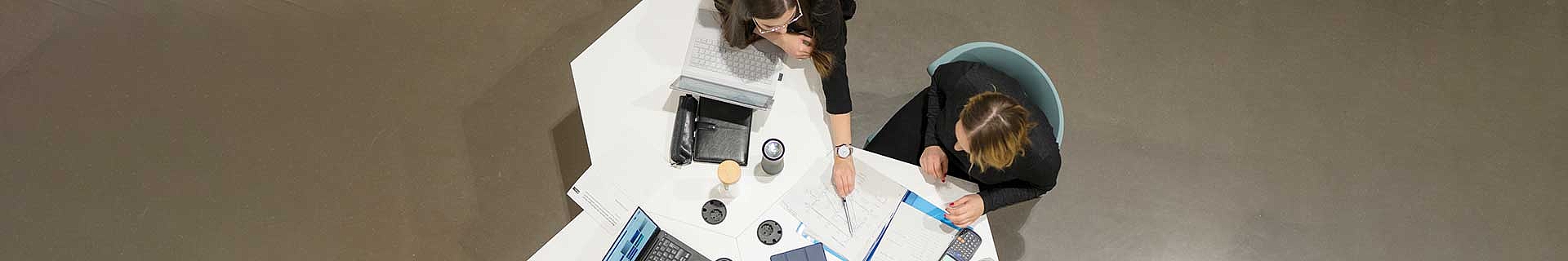 Das Bild zeigt eine Draufsicht auf zwei Personen, die an einem weißen Tisch sitzen. Eine Person arbeitet an einem Laptop, während die andere mit einem Stift auf ein Dokument zeigt. Auf dem Tisch befinden sich verschiedene Gegenstände: ein weiterer Laptop, Notizbücher, Papiere und Tassen. Es sind keine mathematischen Aufgaben oder Hausaufgaben erkennbar – die Szene wirkt wie ein Arbeits- oder Besprechungsumfeld.