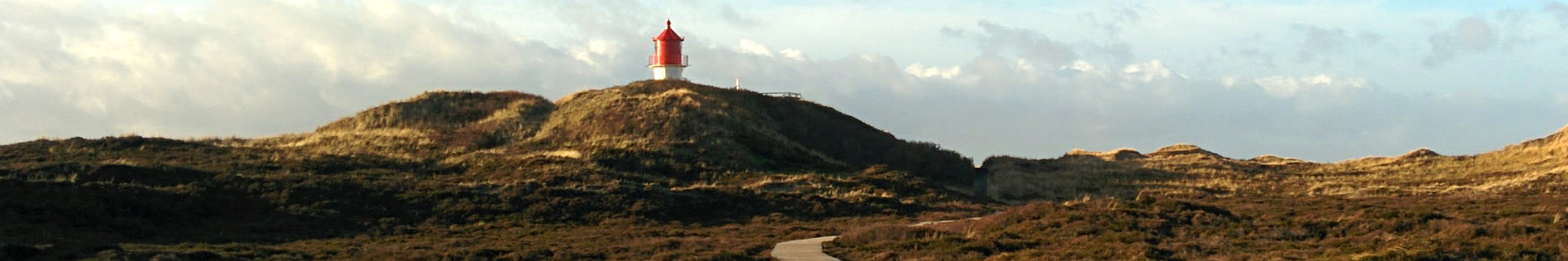 Das Bild zeigt eine malerische Landschaft mit einem Leuchtturm, der auf einem Hügel steht. Der Leuchtturm hat ein rotes Dach und weiße Wände. Umgeben ist er von grünen, grasbewachsenen Hügeln und natürlicher Vegetation. Ein gewundener Pfad führt den Hügel hinauf zum Leuchtturm. Der Himmel ist teilweise bewölkt und trägt zur stimmungsvollen Atmosphäre des Bildes bei.