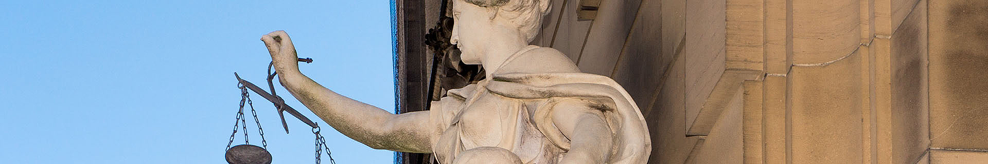 Das Bild zeigt eine Nahaufnahme der Statue der Justitia (Lady Justice). Sie hält in ihrer linken Hand eine Waage, die für Gerechtigkeit und Ausgewogenheit steht. Im Hintergrund ist ein Teil eines Gebäudes mit architektonischen Details sowie ein klarer blauer Himmel zu sehen. Die Darstellung ist ein klassisches Symbol für das Rechtssystem. 