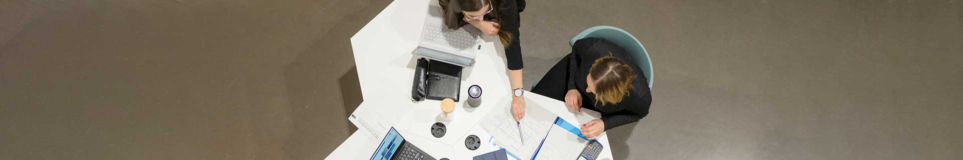 Das Bild zeigt eine Draufsicht auf zwei Personen, die an einem weißen Tisch sitzen. Eine Person arbeitet an einem Laptop, während die andere mit einem Stift auf ein Dokument zeigt. Auf dem Tisch befinden sich verschiedene Gegenstände: ein weiterer Laptop, Notizbücher, Papiere und Tassen. Es sind keine mathematischen Aufgaben oder Hausaufgaben erkennbar – die Szene wirkt wie ein Arbeits- oder Besprechungsumfeld.